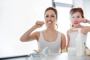 Waist up portrait of woman with toothbrush and her pretty child cleaning teeth together. They are looking concentrated and high-spirited
