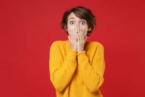 Shocked amazed surprised excited young brunette woman 20s wearing basic casual yellow sweater stand covering mouth with hands looking camera isolated on bright red colour background studio portrait