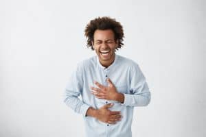 Positive and joyful Afro American male with fine crop of hair bursting into laughing holding his hands on stomach can`t stopping laughing after hearing funny anecdote. Positive emotions and humour