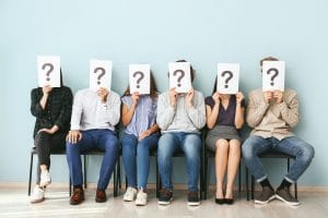 Young people hiding faces behind paper sheets with question marks while waiting for job interview indoors