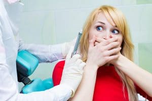 Closeup portrait terrified girl woman scared of needles, syringes, dentist visit siting in chair, opened mouth doesn't want dental procedure drilling tooth extraction isolated clinic office background