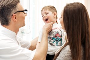 Dentist examining teeth of little boy with his mother watching at dental clinic