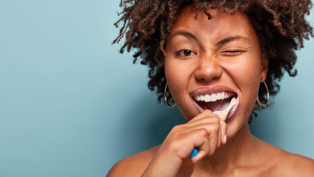 Tooth care concept. Slim naked young woman blinks eye, has daily routine after shower, brushes teeth with tootbrush, has healthy skin, isolated over blue background, free space for your promotion.