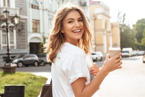 Portrait of smiling european woman strolling through city street with silver laptop and takeaway coffee in hands
