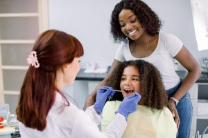 Dental treatment without fear. Female dentist in whilte uniform and gloves, examining teeth of little African American girl, while her mother supporting her behind, at modern pediatric dental clinic.