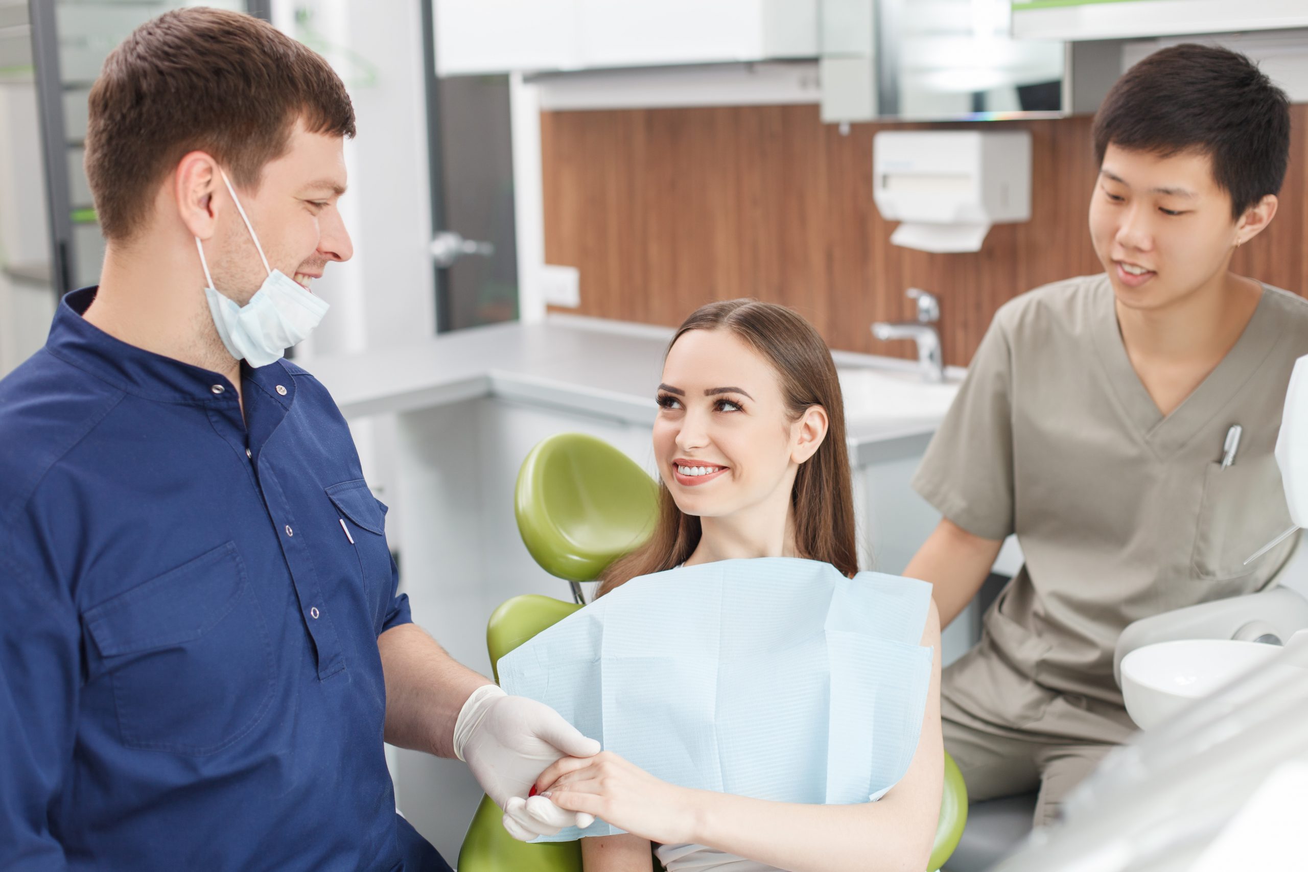 Cheerful dentist and his assistant are calming their visitor. The dentist is holding female hand with support. The woman is sitting in medical chair and looking at dentist with trust. They are smiling