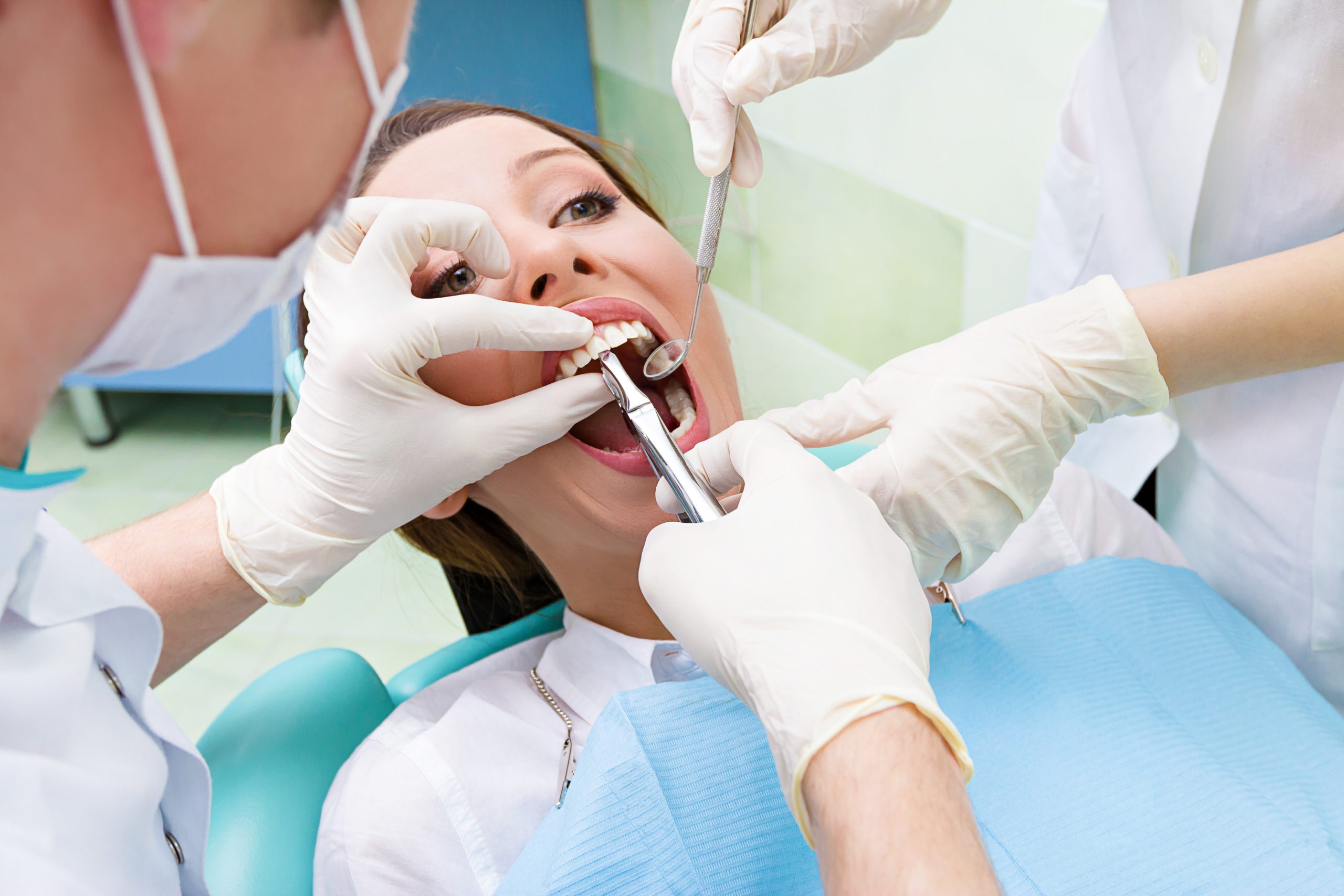 Closeup portrait dentist, his assistant carrying out thorough examination dental procedure, tooth extraction on young female patient, lying in chair, wide open mouth isolated clinic office background