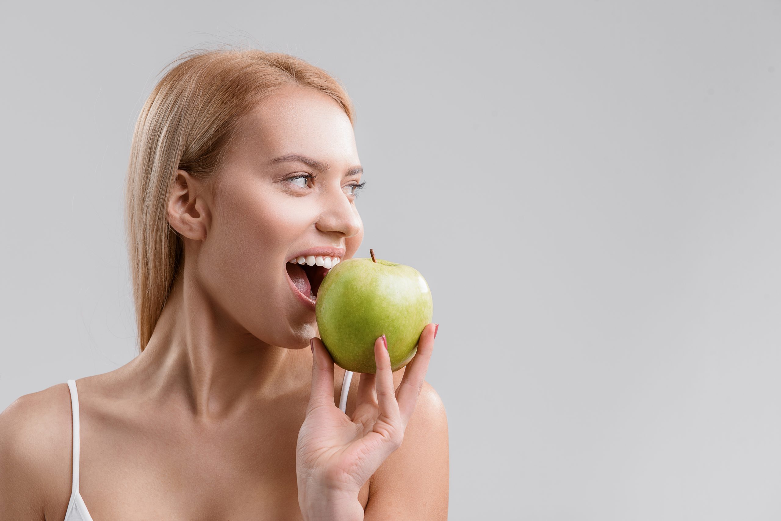 Happy young woman is biting green apple with appetite. She is standing and looking aside with joy. Isolated and copy space in right side