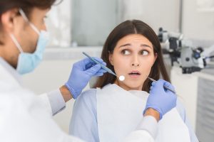Scared young woman looking at dentist with eyes wide open, doctor holding tools, fear of dentistry concept