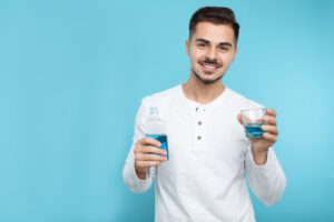 Young man holding bottle and glass with mouthwash on color background, space for text. Teeth and oral care