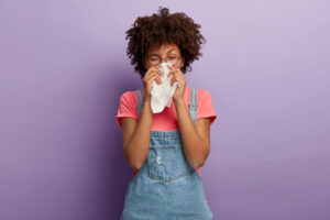 Portrait of sick African American woman sneezes in white tissue, suffers from rhinitis and running nose, has allergy on something, looks unhealthy, feels unwell. Symptoms of cold or allergy.