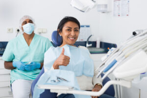 Happy latin american woman sitting in dental chair after teeth cure in modern clinic, gesturing thumbs up