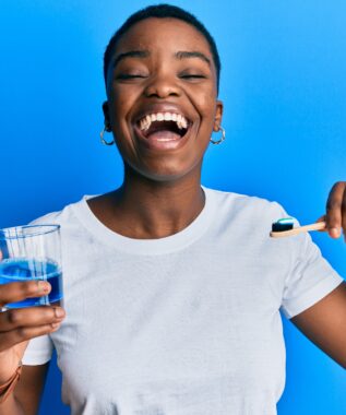 Young african american woman holding glass of mouthwash and toothbrush for fresh breath smiling and laughing hard out loud because funny crazy joke.