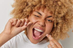 Close up shot of curly haired woman removes food uses thread or dental floss winks eye keeps mouth opened takes care of her mouth cavity poses indoor. Teeth flossing and oral hygiene concept.