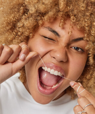 Close up shot of curly haired woman removes food uses thread or dental floss winks eye keeps mouth opened takes care of her mouth cavity poses indoor. Teeth flossing and oral hygiene concept.