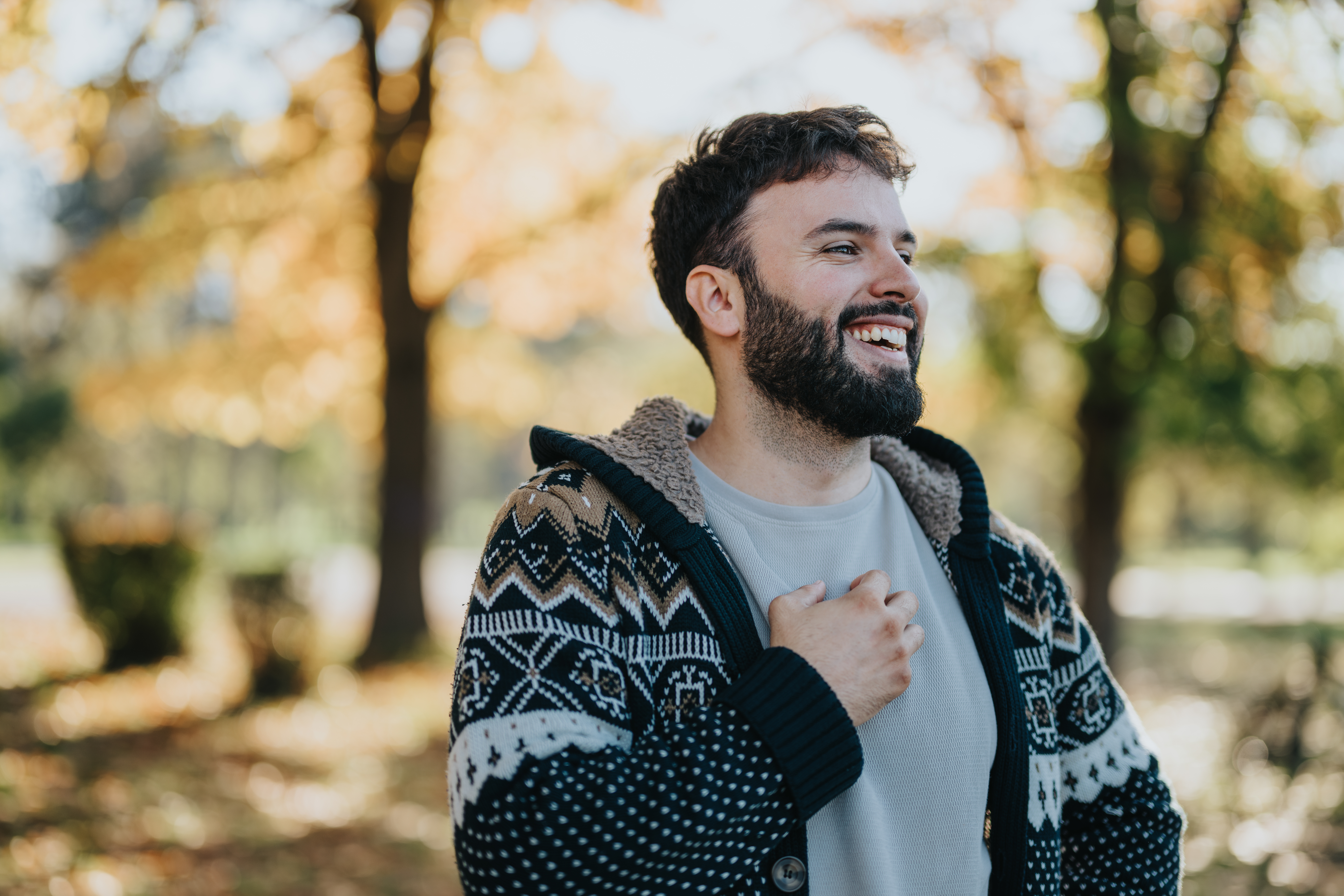 A cheerful man in a warm, patterned sweater stands amidst autumn trees, laughing freely. The sunlit background and fallen leaves capture the essence of an enjoyable, relaxed outdoor moment. sheridan wy broken tooth